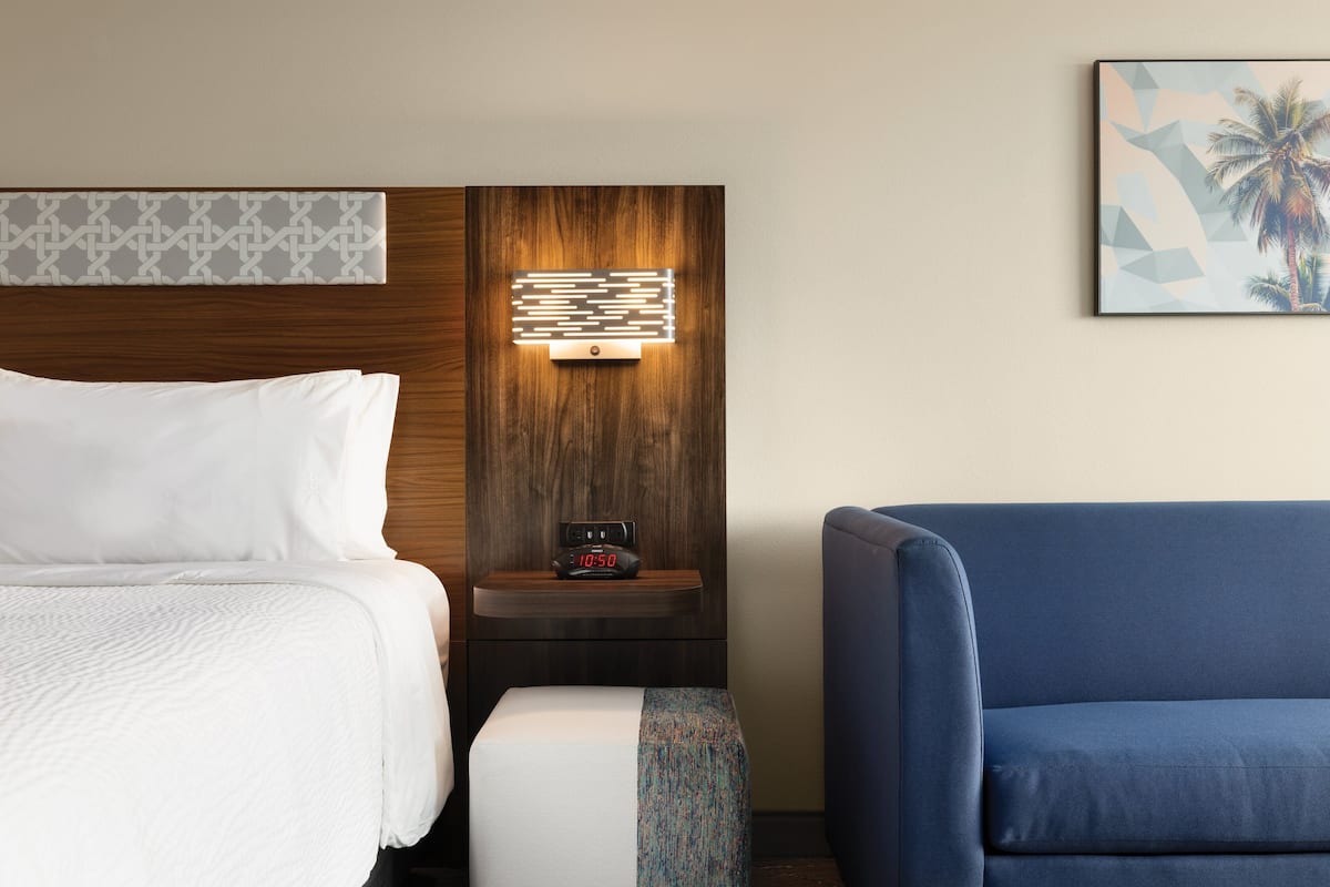 Une chambre d’hôtel avec un lit, une table de chevet avec une horloge, un fauteuil bleu et une œuvre d’art accrochée au mur.
