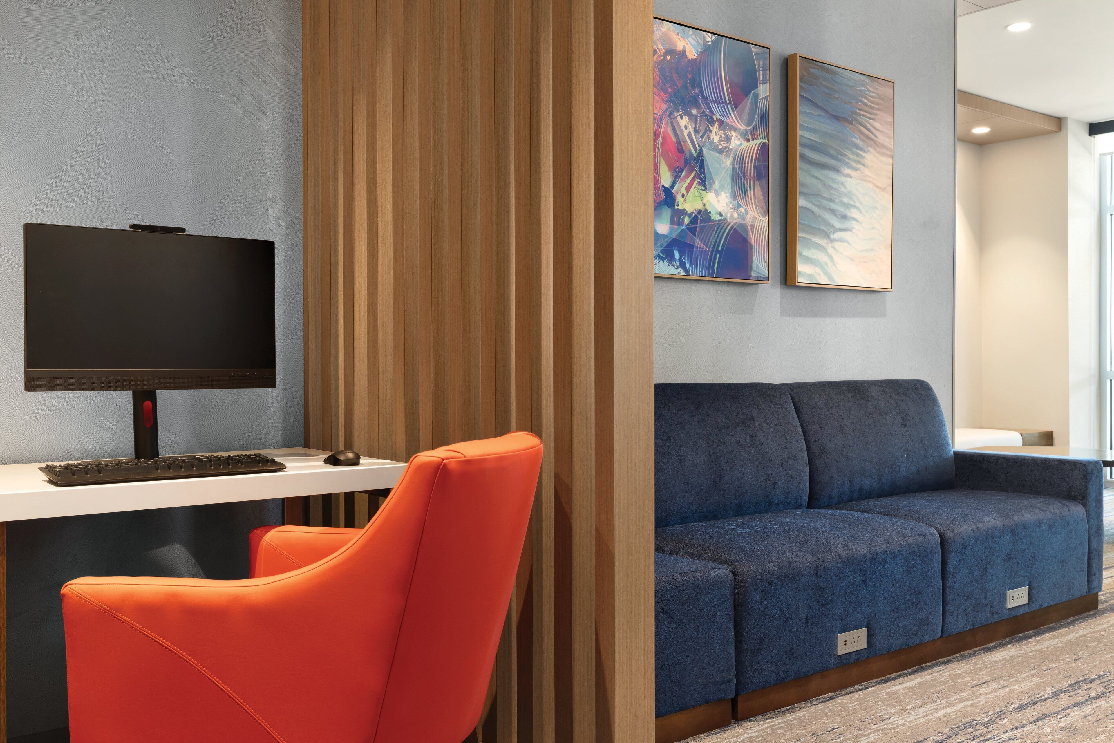 A modern hotel lobby with a flat-screen TV, a blue sofa, an orange chair, and abstract art on the wall.