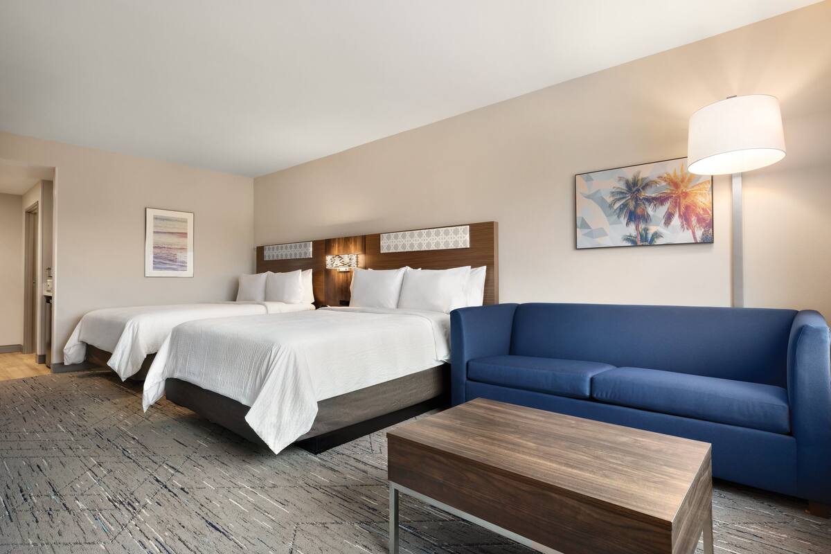 Habitación de hotel con dos camas, un sofá azul, una mesa de centro de madera y un cuadro de palmeras.