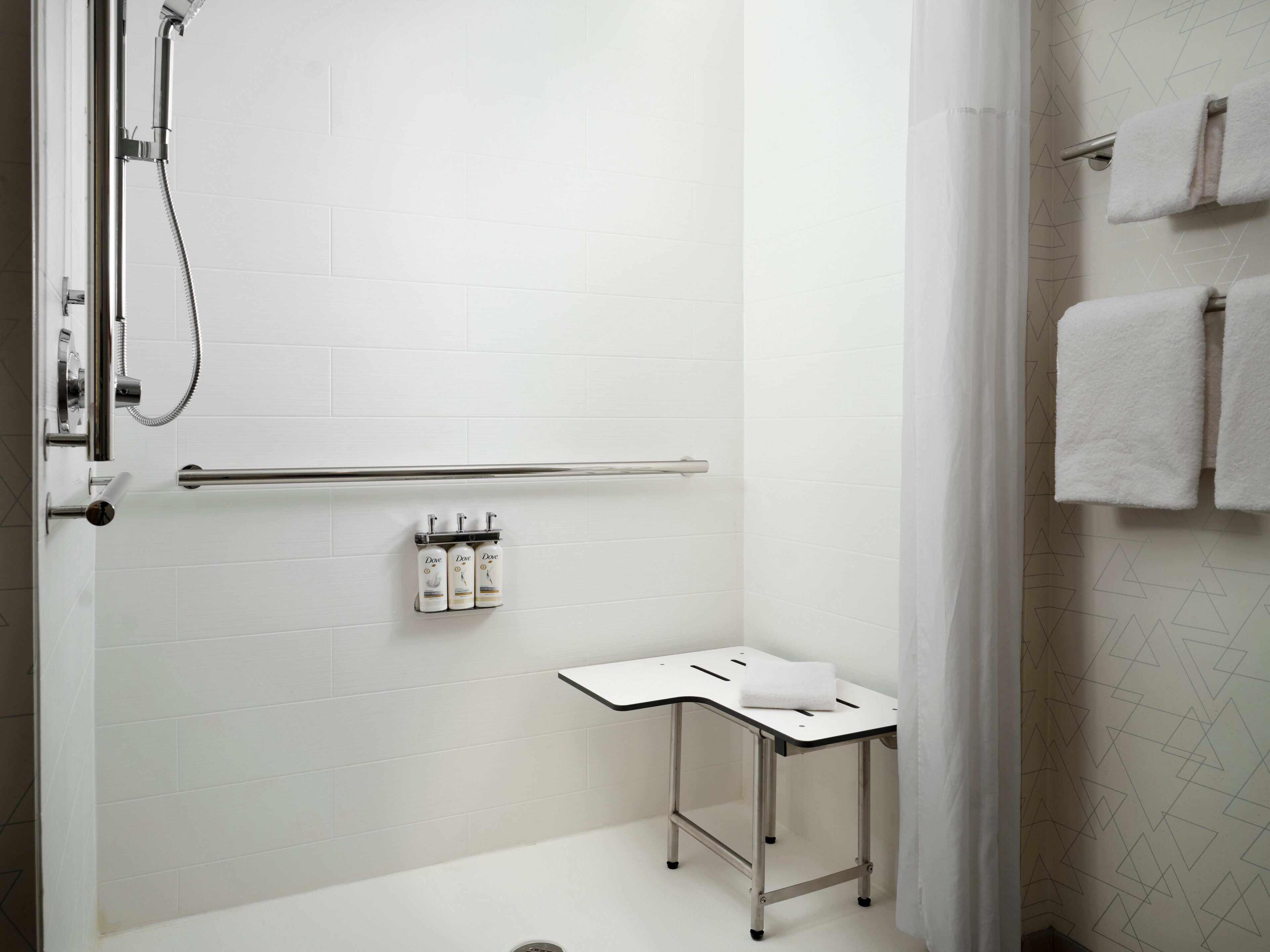 Een moderne badkamer met witte tegels, een douche met handdouche, een handdoekrek met twee handdoeken en een klein tafeltje met een handdoek.