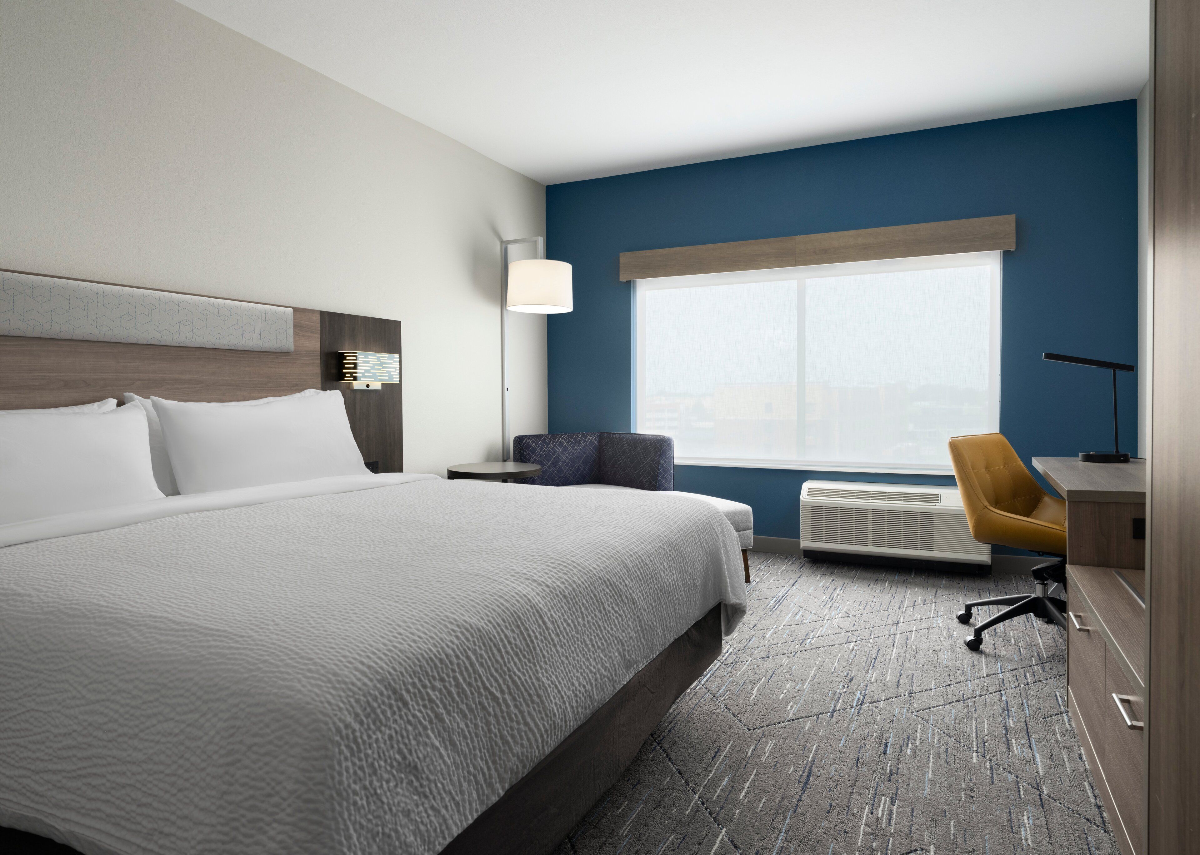 A hotel room with a large bed, a desk with a chair, a window with blinds, and a wall-mounted air conditioner.