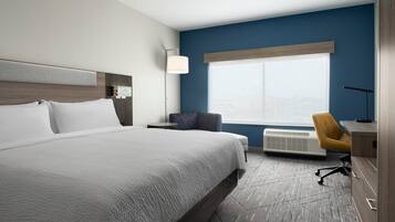 A hotel room with a large bed, a desk with a chair, a window with blinds, and a wall-mounted air conditioner.