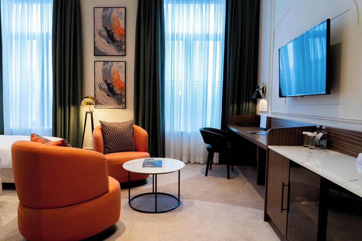 A modern hotel room with a sofa, a round coffee table, a desk, and a flat-screen TV mounted on the wall.
