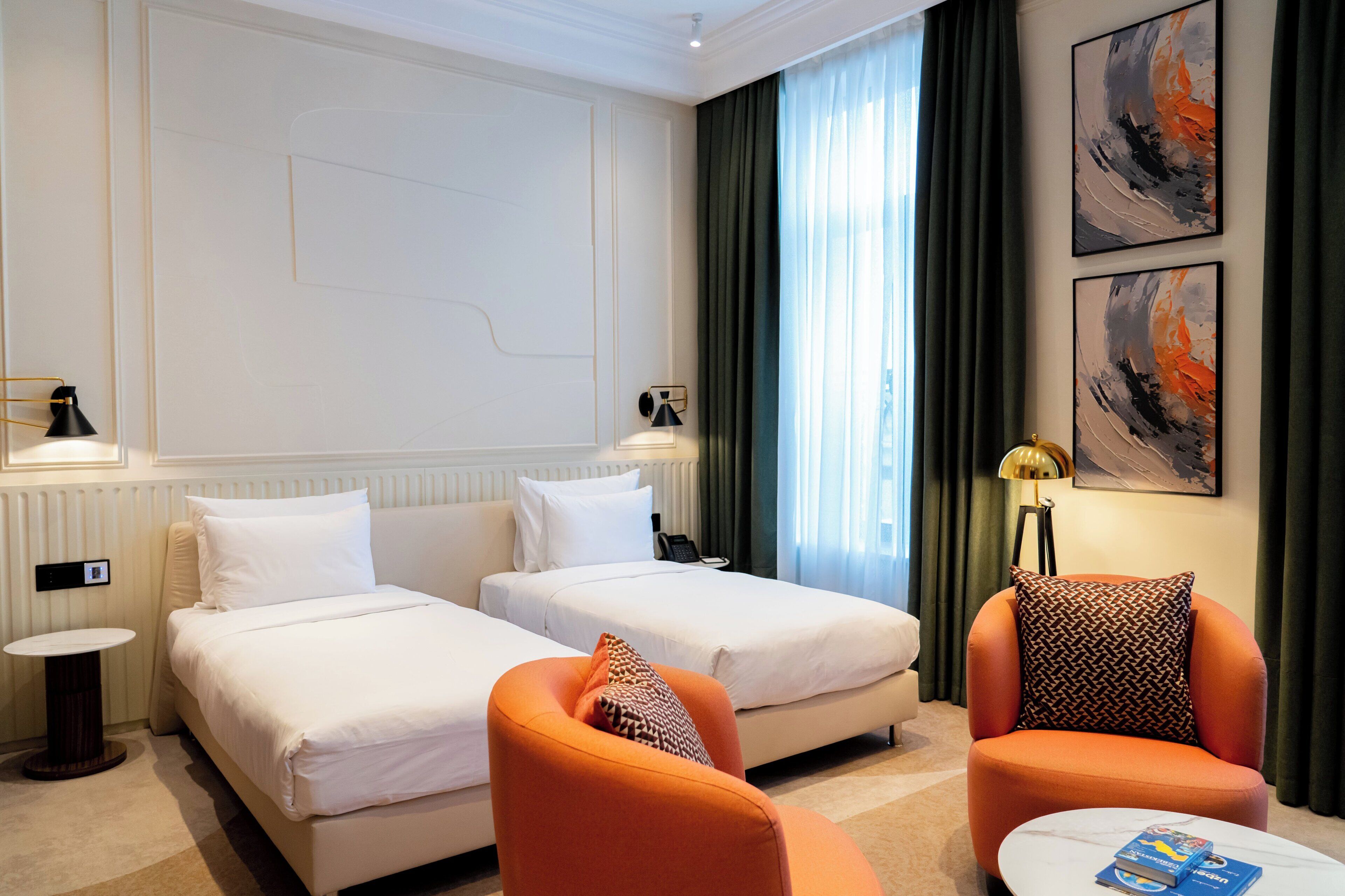 A hotel room with two beds, a small table, an orange armchair, a round coffee table, and two abstract paintings on the wall.