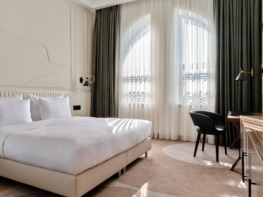 A hotel room with a large bed, a desk with a chair, a window with curtains, and a radiator on the wall.