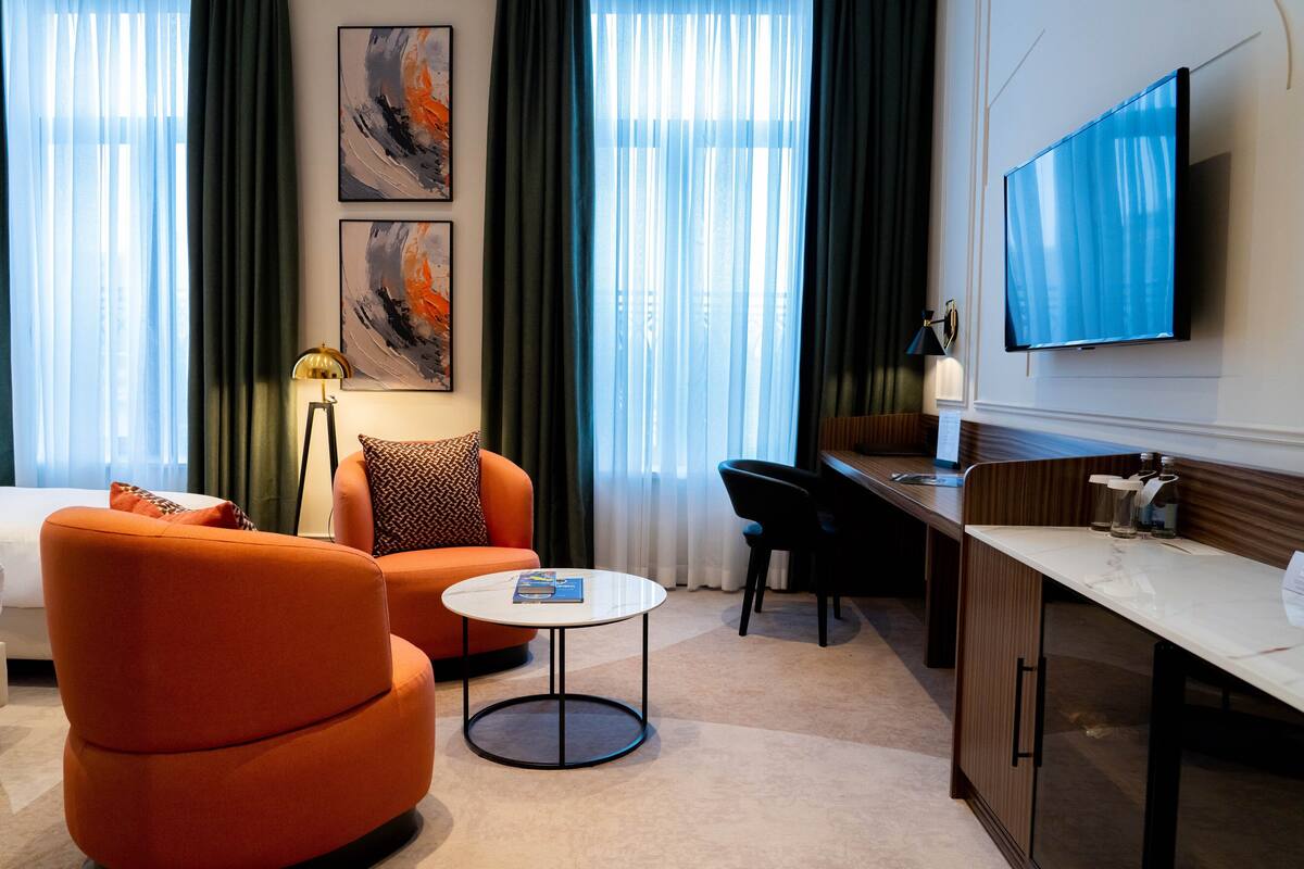 A modern hotel room with a sofa, a round coffee table, a desk, and a flat-screen TV mounted on the wall.