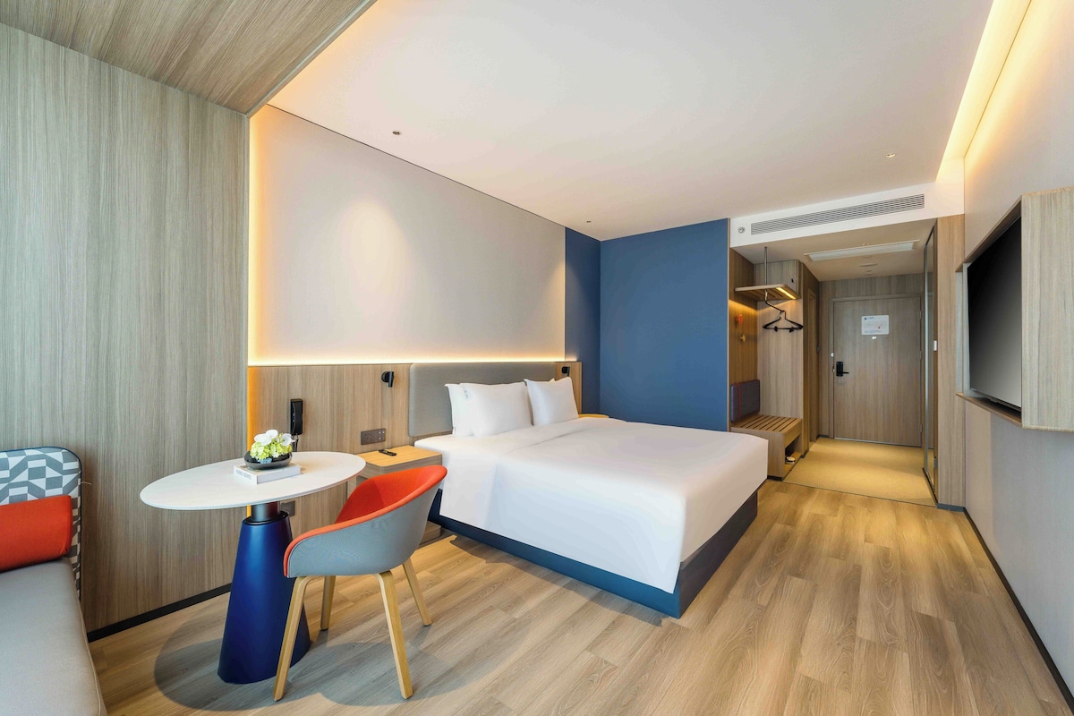 A modern hotel room with a large bed, a small round table, a chair, and a TV mounted on the wall.