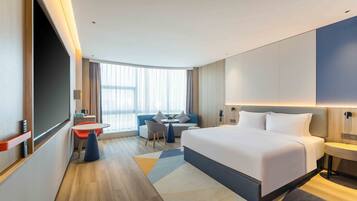 Quarto de hotel moderno com uma cama grande, área de estar com mesa e uma janela ampla que permite a entrada de luz natural.