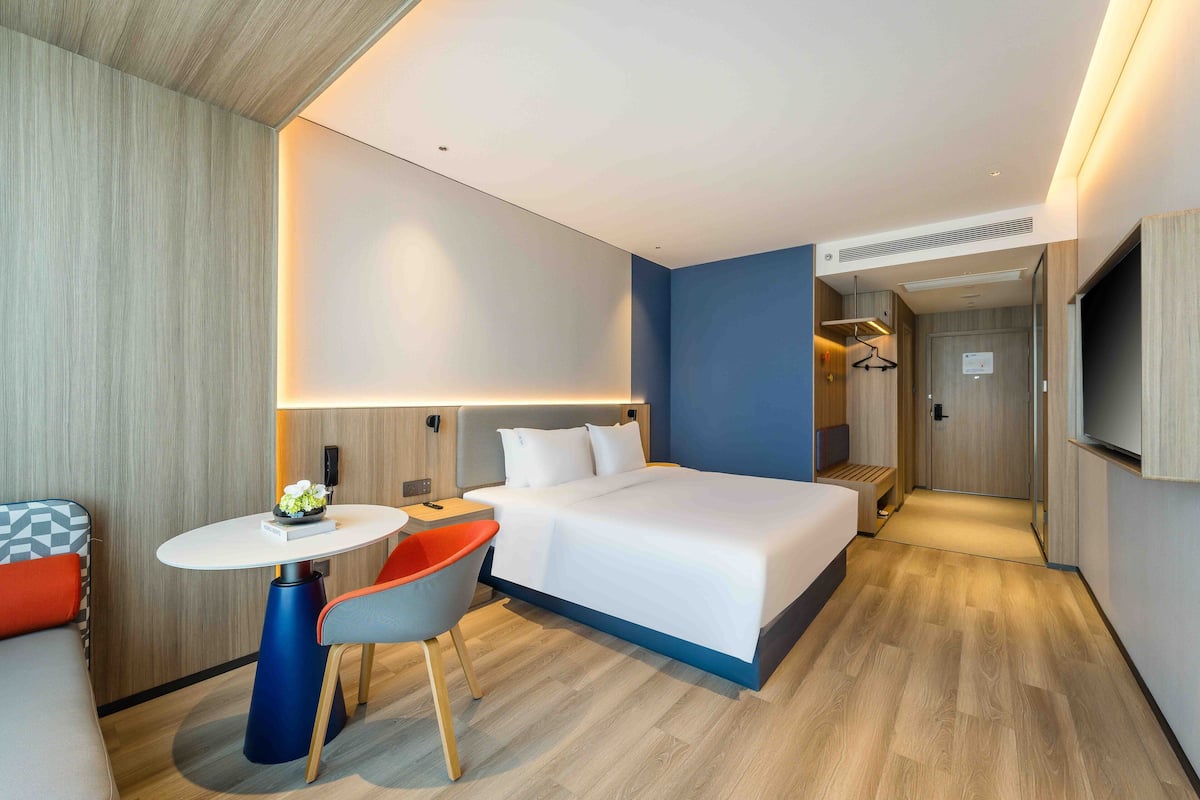 A modern hotel room with a large bed, a small round table, a chair, and a TV mounted on the wall.