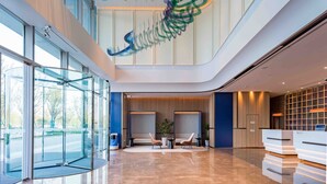 Un lobby moderno de hotel con entrada de vidrio, un mostrador de recepción, zona de estar y un techo con una lámpara de diseño único.