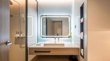 Un baño moderno con cabina de ducha de vidrio, un espejo grande con iluminación integrada, un lavabo blanco y muebles de madera.