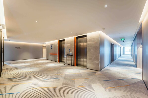 A modern hotel corridor with a patterned carpet, elevators, and a long hallway.