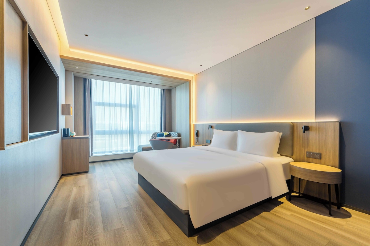 A modern hotel room with a large bed, bedside table, and a view of the city through large windows.
