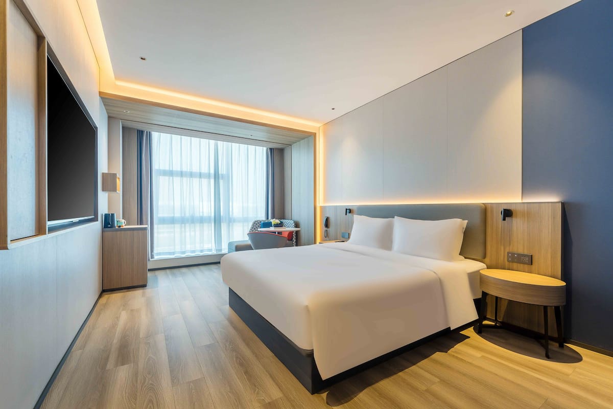 A modern hotel room with a large bed, bedside table, and a view of the city through large windows.