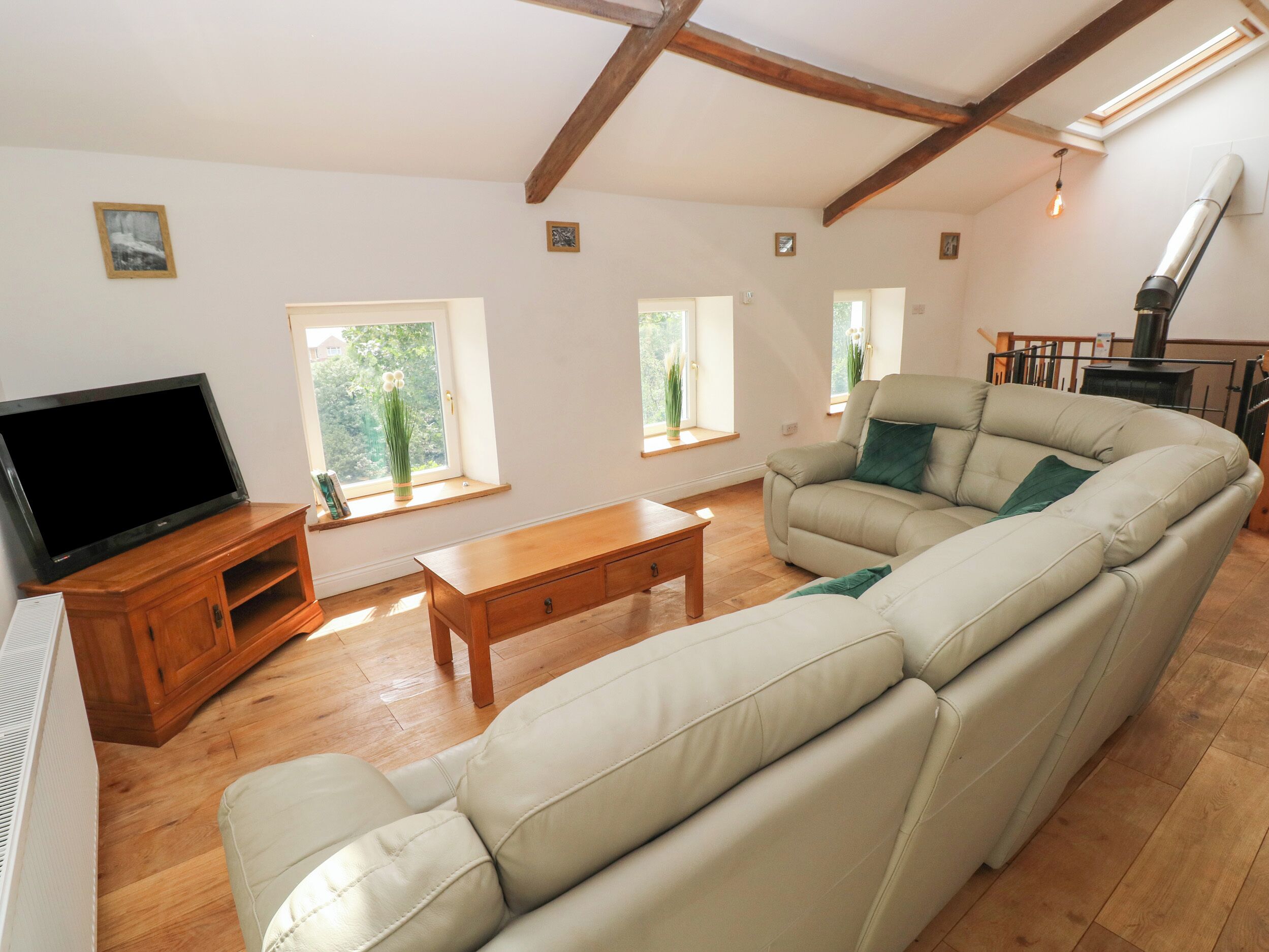 A spacious living room with a large sofa, a wooden coffee table, and a flat-screen TV on a wooden stand. There are windows allowing natural light and a view of greenery outside.