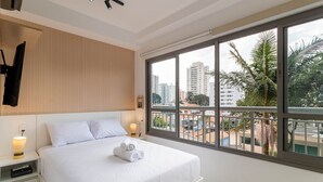 A hotel room with a large bed, a nightstand with a lamp, and a window with a city view. - Studio ID Lisboa (São Paulo)