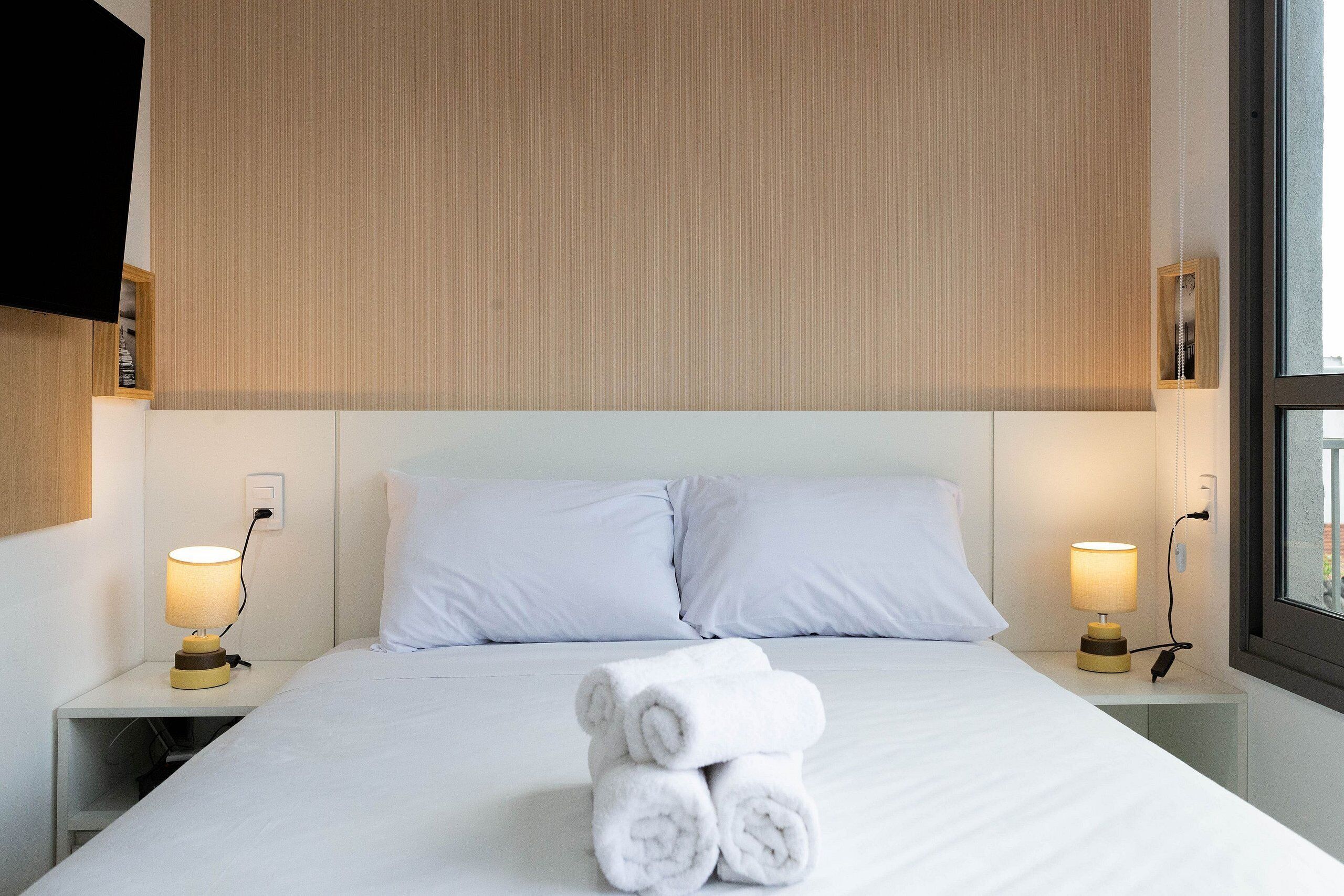 Una cama bien hecha con sábanas blancas y toallas enrolladas, dos lámparas de noche y un cabecero de madera.