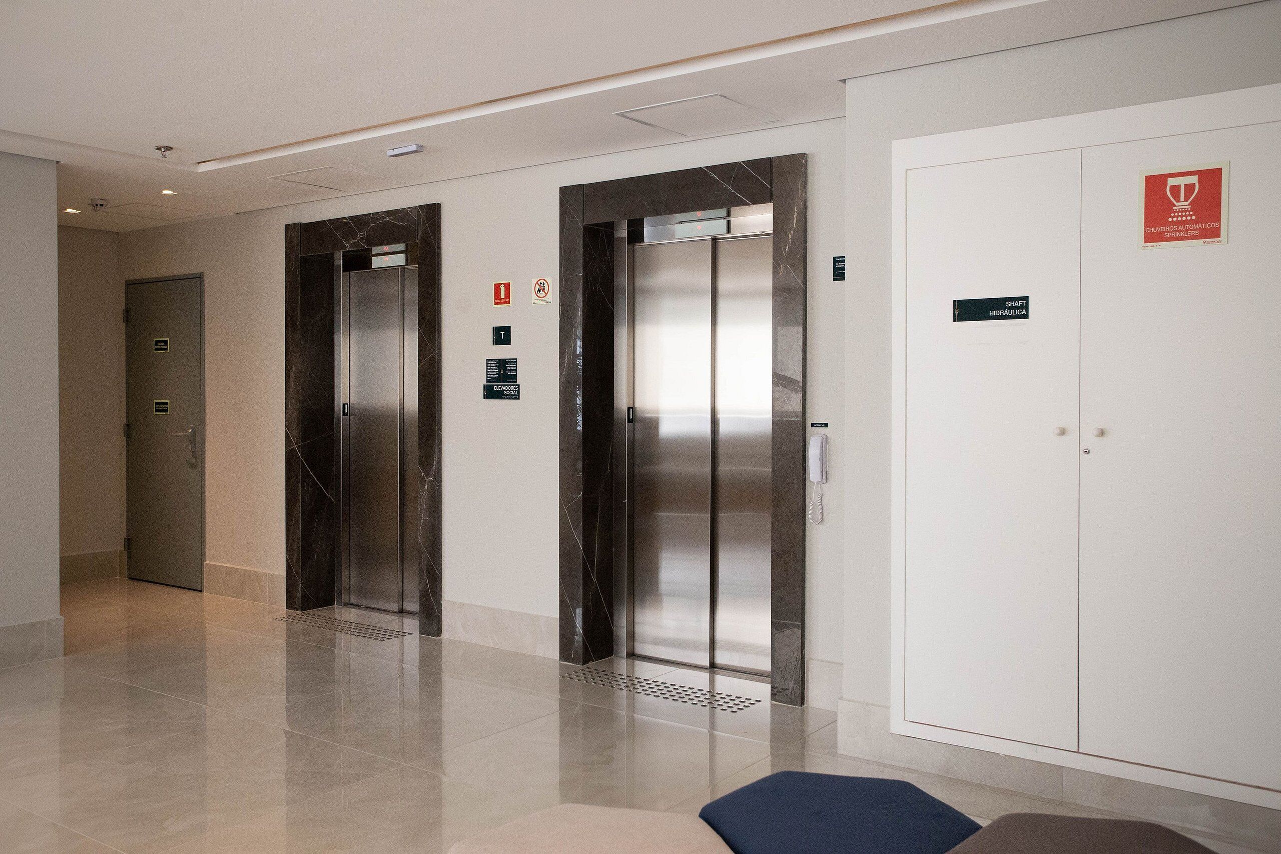 A modern elevator area with two elevators, a white door with a red sign, and a floor with a marble-like finish.