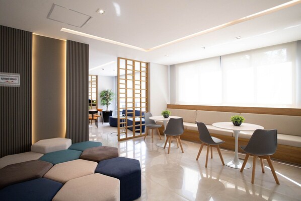 A modern lounge area with seating arrangements, a small table with chairs, and a partition with a sign indicating an elevator service area. - Studio ID Lisboa (São Paulo)