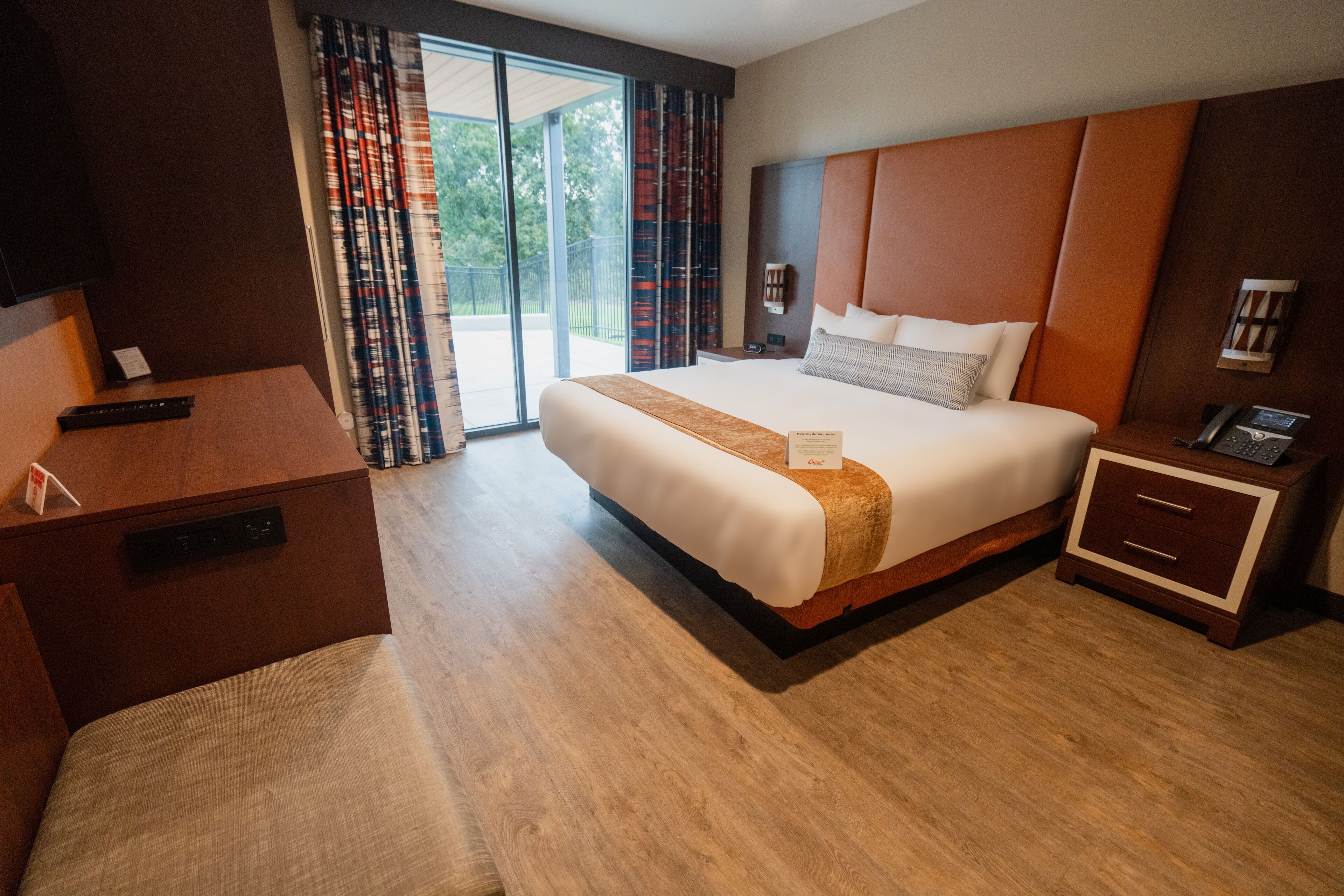A hotel room with a large bed, a desk, a bedside table with a phone, and a view of the outdoors through a sliding door.