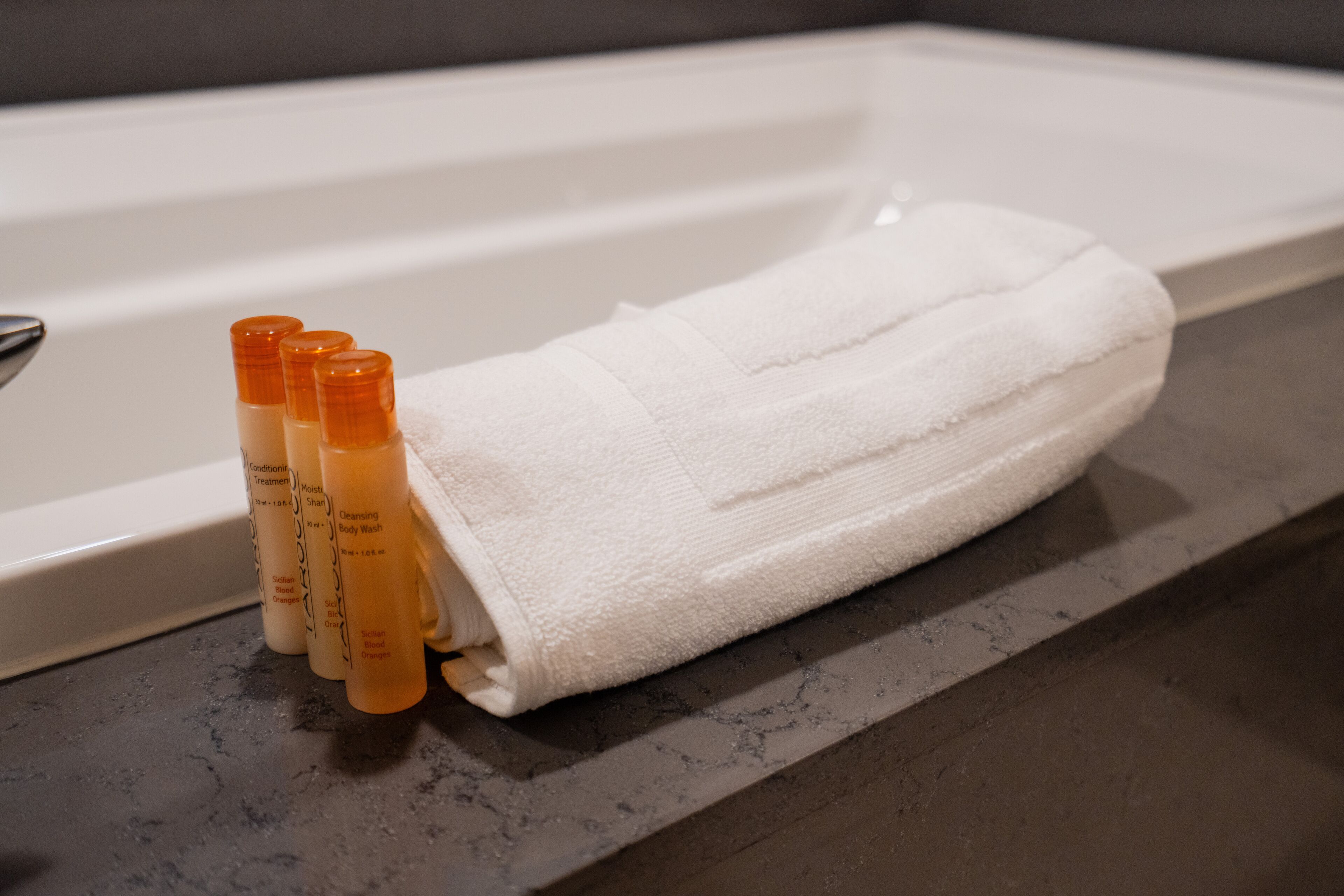 Una alfombra de baño blanca junto a una bañera con tres botellas de jabón de cuerpo.