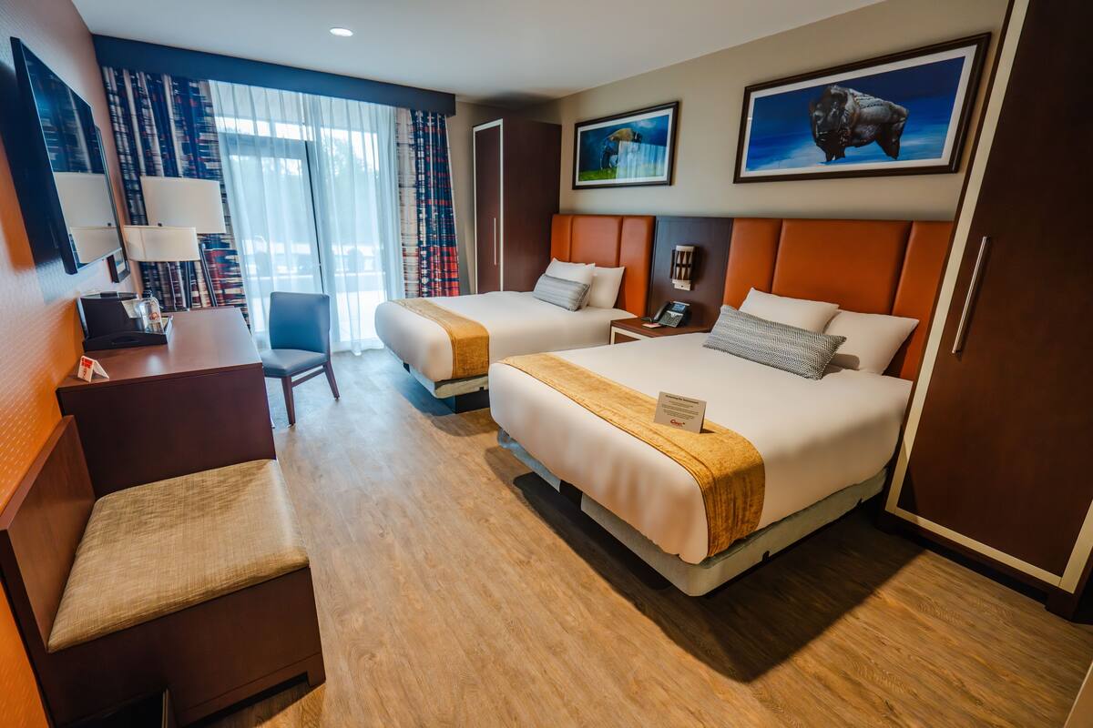 A hotel room with two beds, a desk, a chair, a TV, and a large window with curtains.