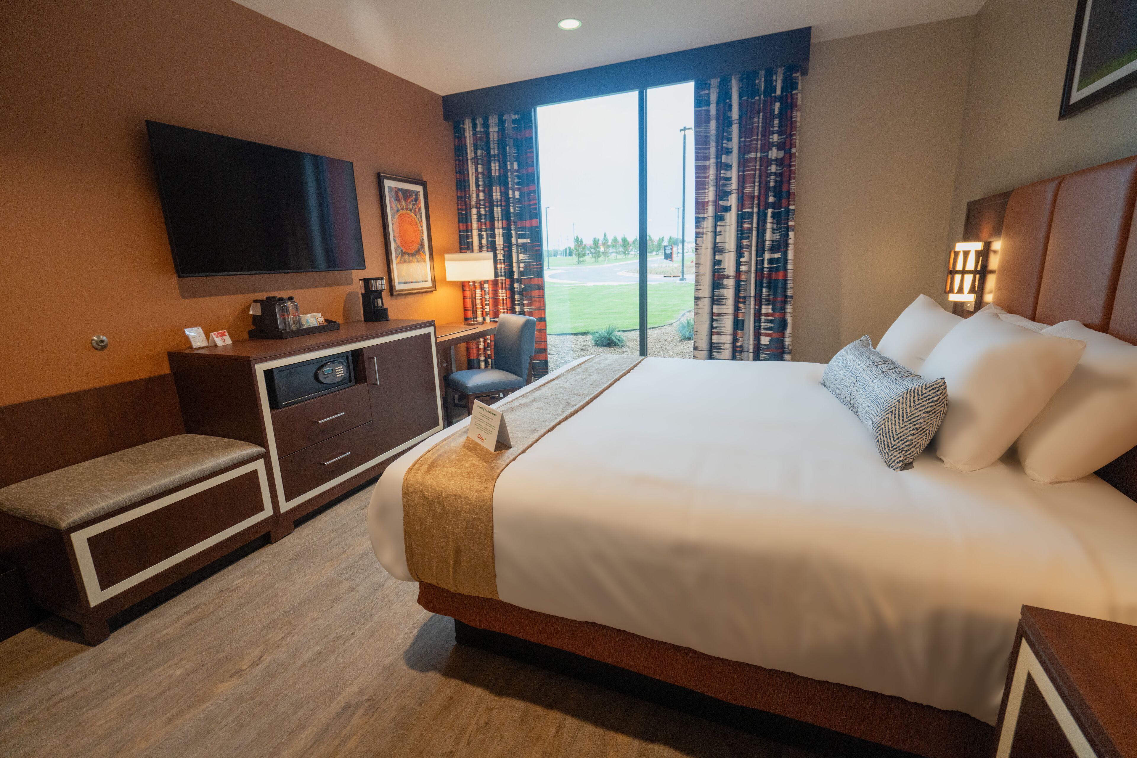 Ein Hotelzimmer mit einem großen Bett, einem Schreibtisch mit Computer, einem an der Wand befestigten Fernseher und Blick ins Freie durch die Fenster.