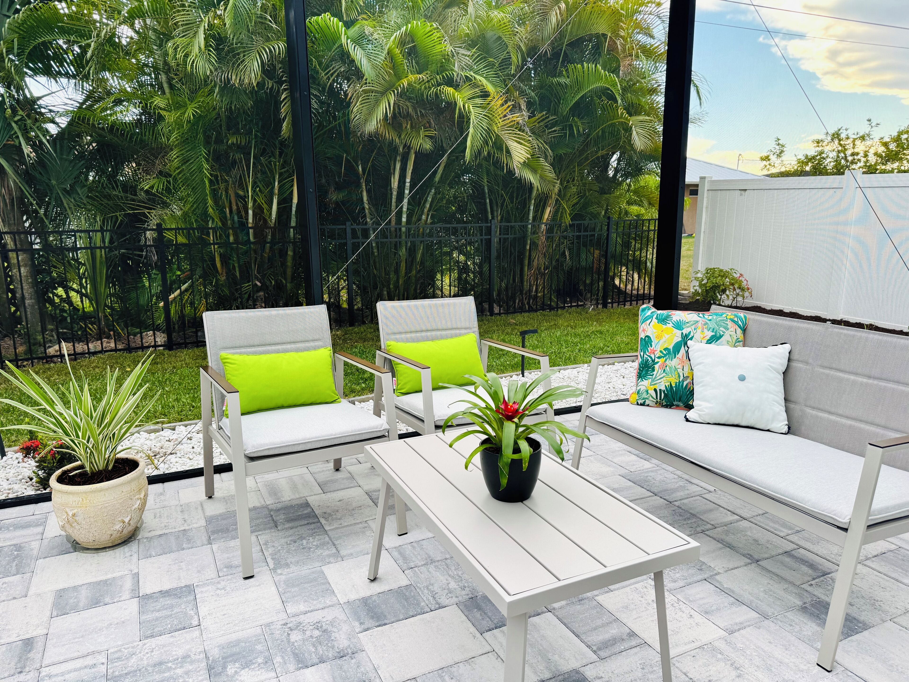 A patio area with a white sofa, two chairs with green cushions, a small table, and potted plants.