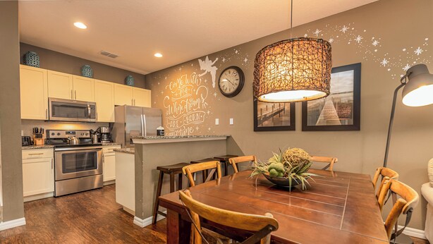 A modern kitchen with stainless steel appliances, a dining area with a wooden table and chairs, and a decorative wall with a clock and a quote. - Spacious 3BR Townhome in Kissimmee, perfect for family getaways! (Kissimmee)