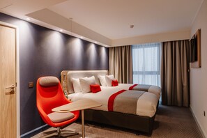 A hotel room with a large bed, a red armchair, a small table, and a window with curtains.