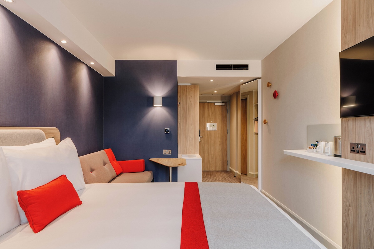 A hotel room with a bed, a small table, a chair, a TV, and a shelf with toiletries.