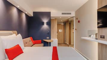 A hotel room with a bed, a small table, a TV, and a wall-mounted light.