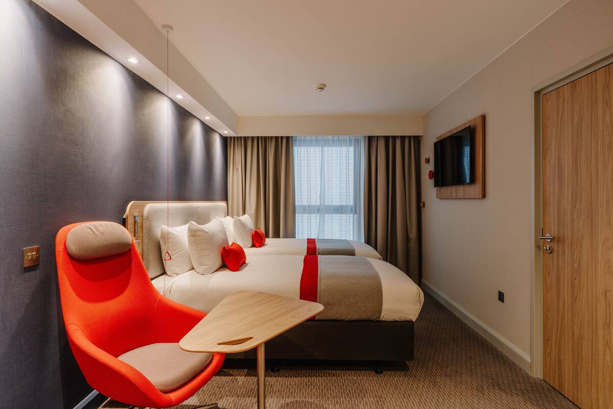 A modern hotel room with a large bed, a bright orange chair, a small wooden table, and a TV mounted on the wall.