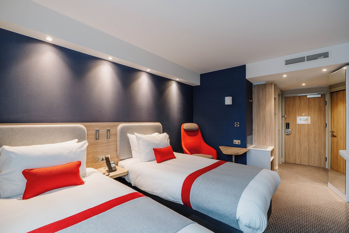 A hotel room with two beds, a red chair, a small table, and a wooden wardrobe.