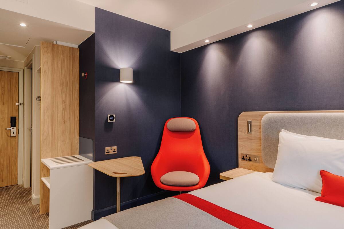 A modern hotel room with a bed, a red chair, a small table, and a wall-mounted light.