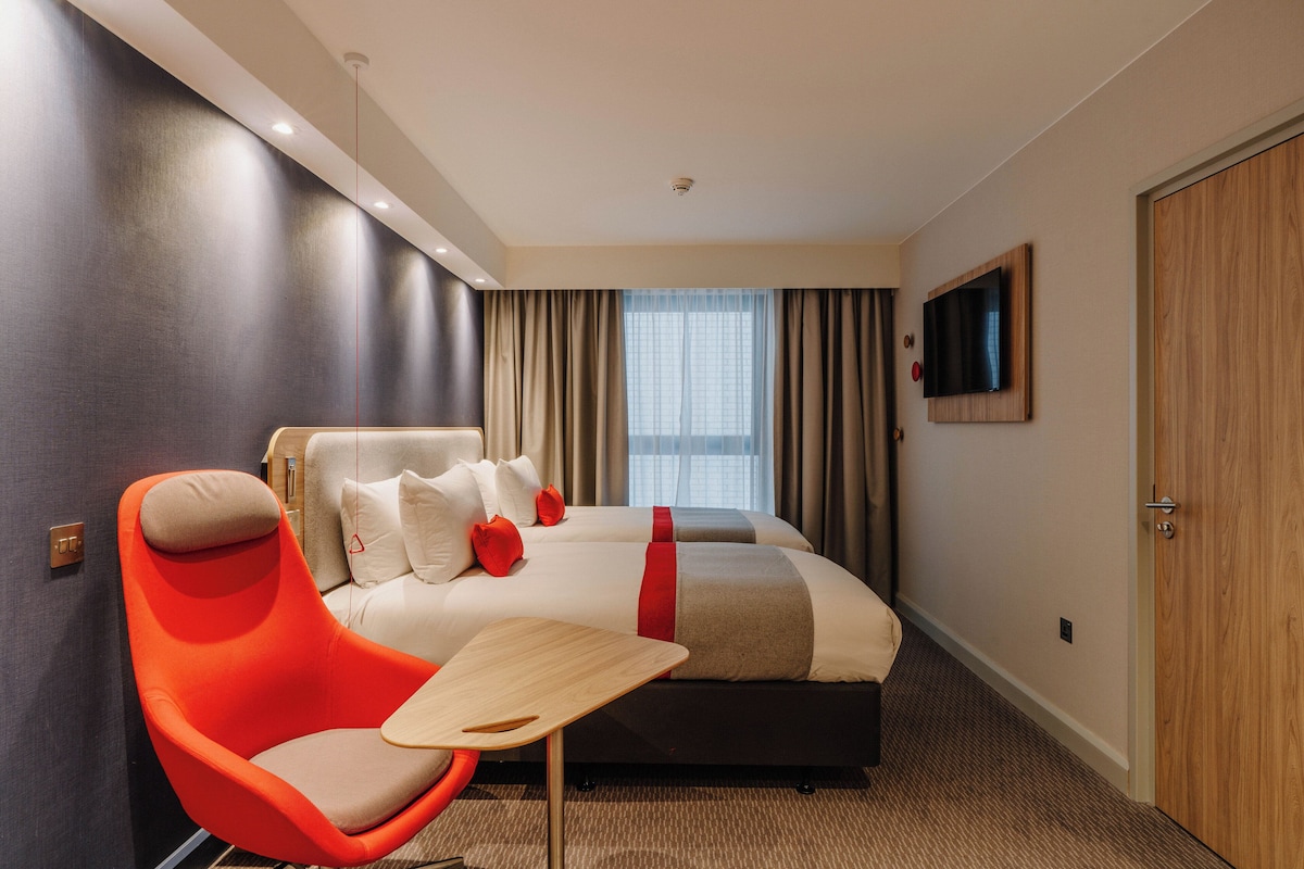 A modern hotel room with a large bed, a bright orange chair, a small wooden table, and a TV mounted on the wall.