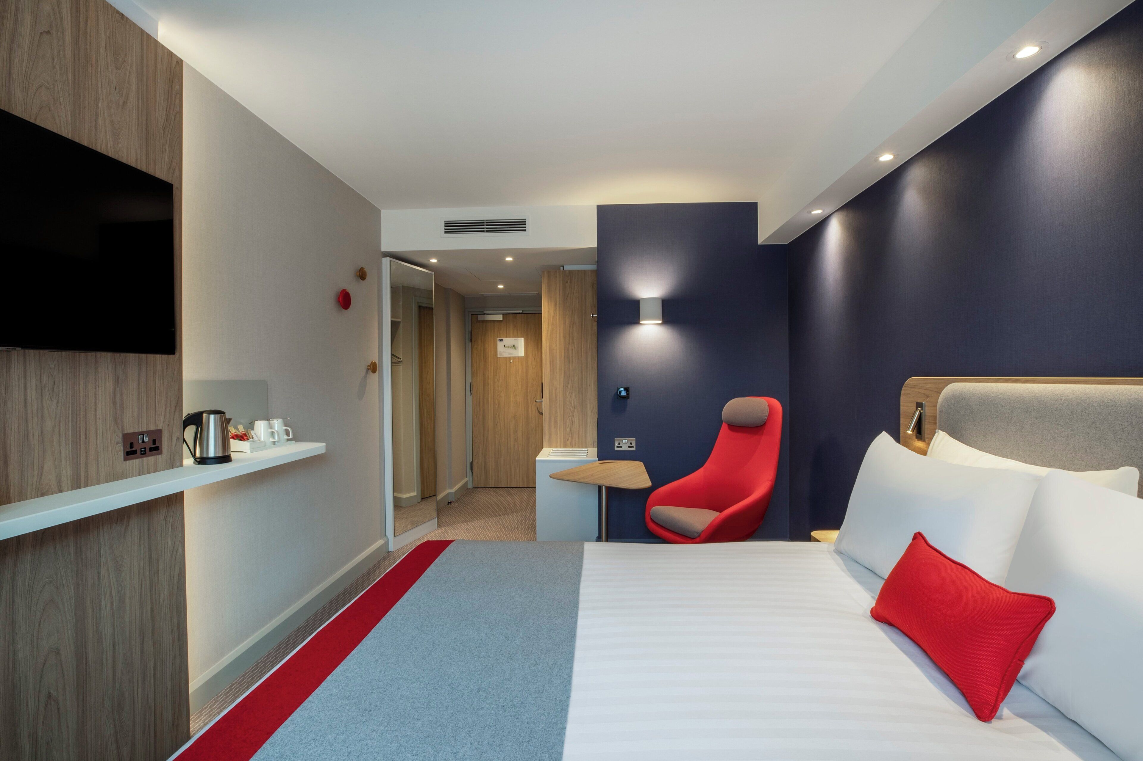 A hotel room with a bed, a red chair, a TV, and a shelf with a kettle and cups.