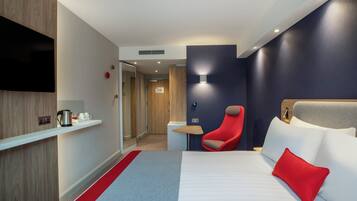 A hotel room with a bed, a red chair, a TV, and a shelf with a kettle and cups.