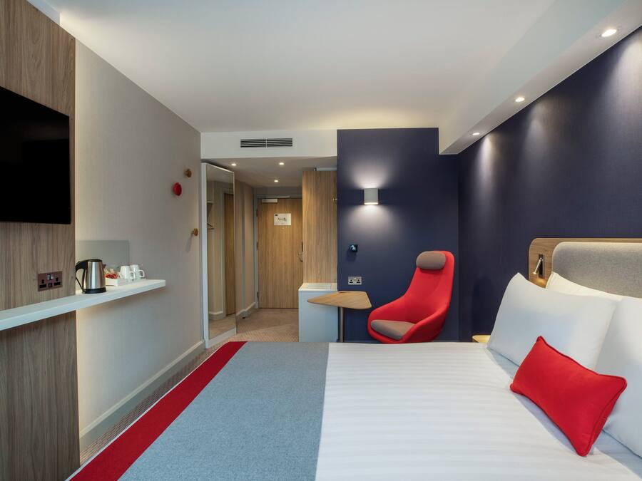 A hotel room with a bed, a red chair, a TV, and a shelf with a kettle and cups.