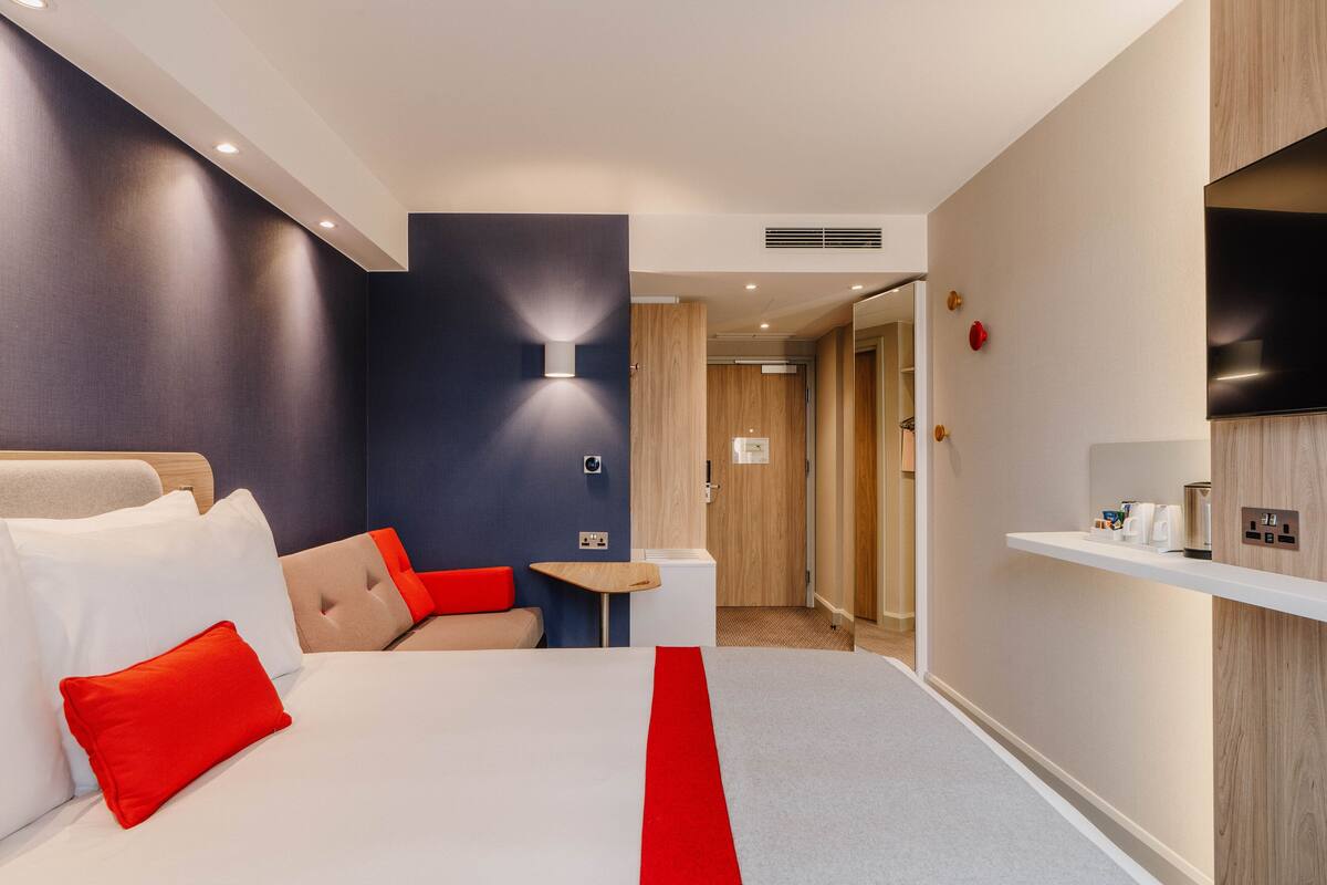 A hotel room with a bed, a small table, a chair, a TV, and a shelf with toiletries.