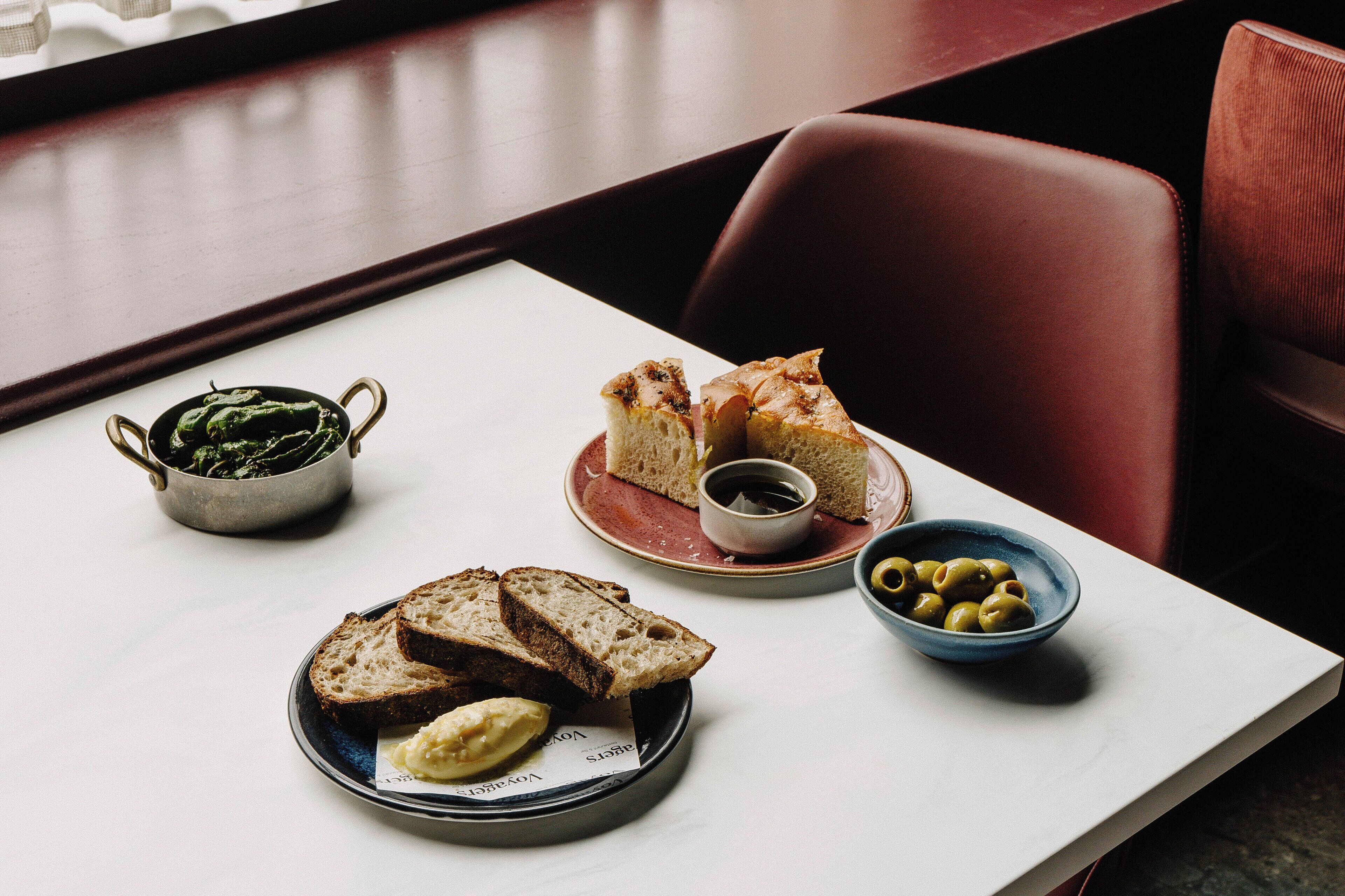 Una mesa con una fuente de pan, un cuenco de aceitunas y un bol de verduras.