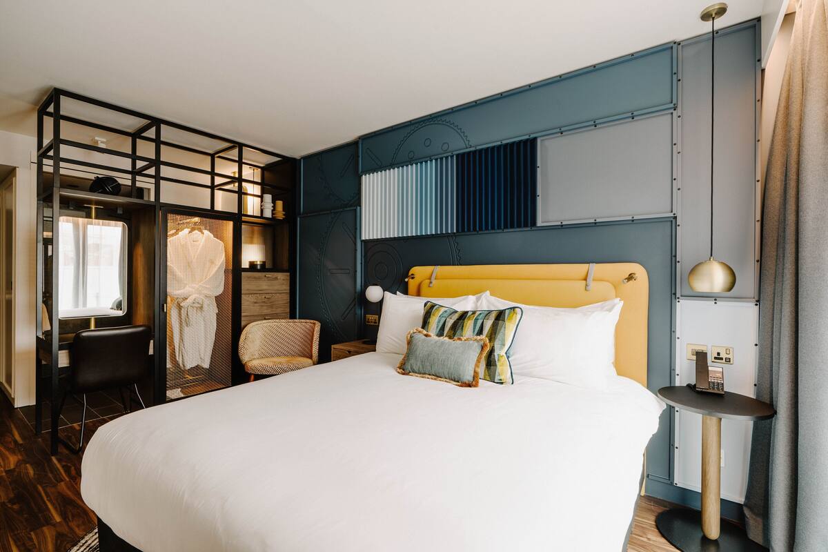 A modern hotel room with a large bed, a desk, a chair, and a wardrobe.