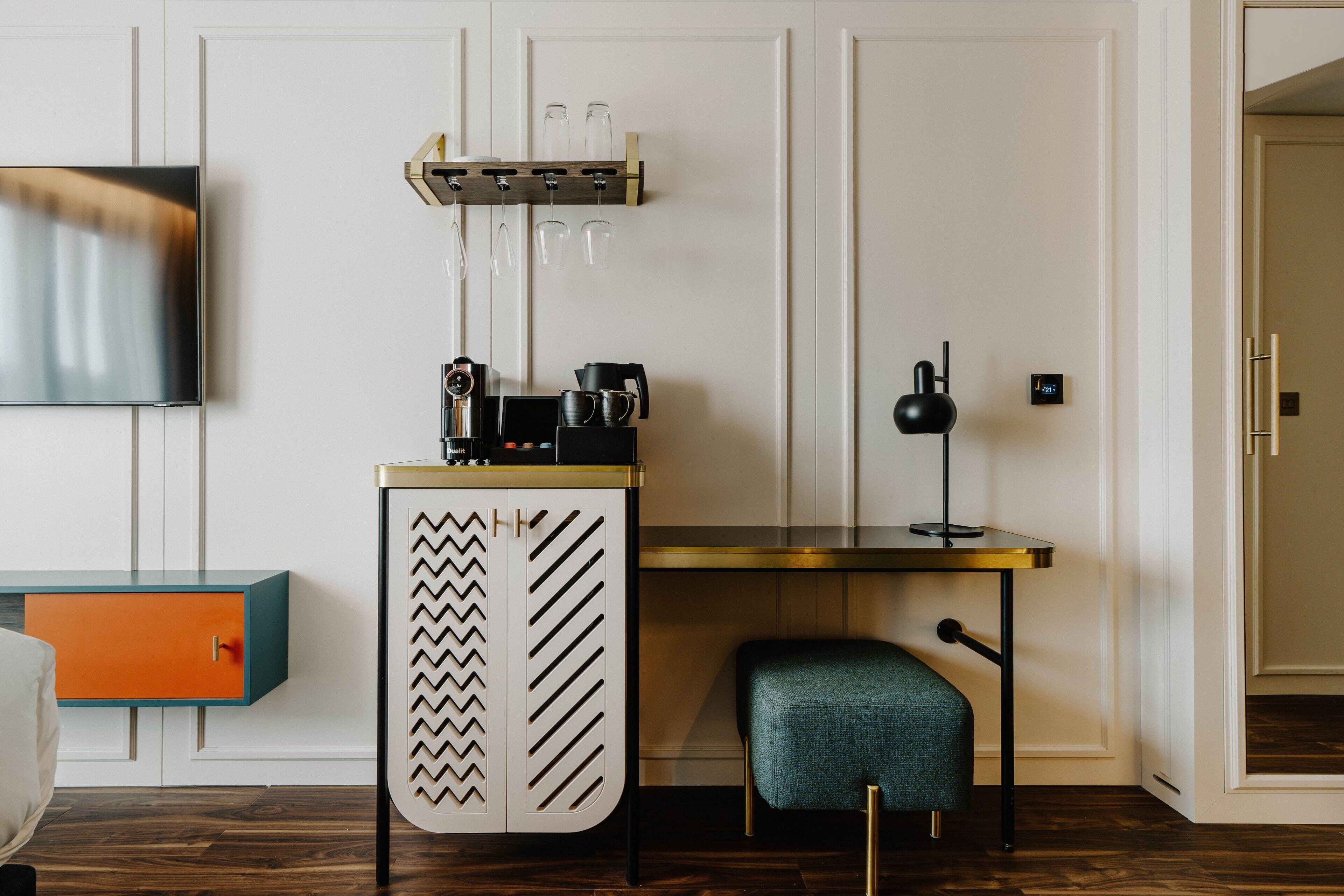 A compact room with a wooden floor, a small desk with a chair, a television mounted on the wall, and a cabinet with a unique design.