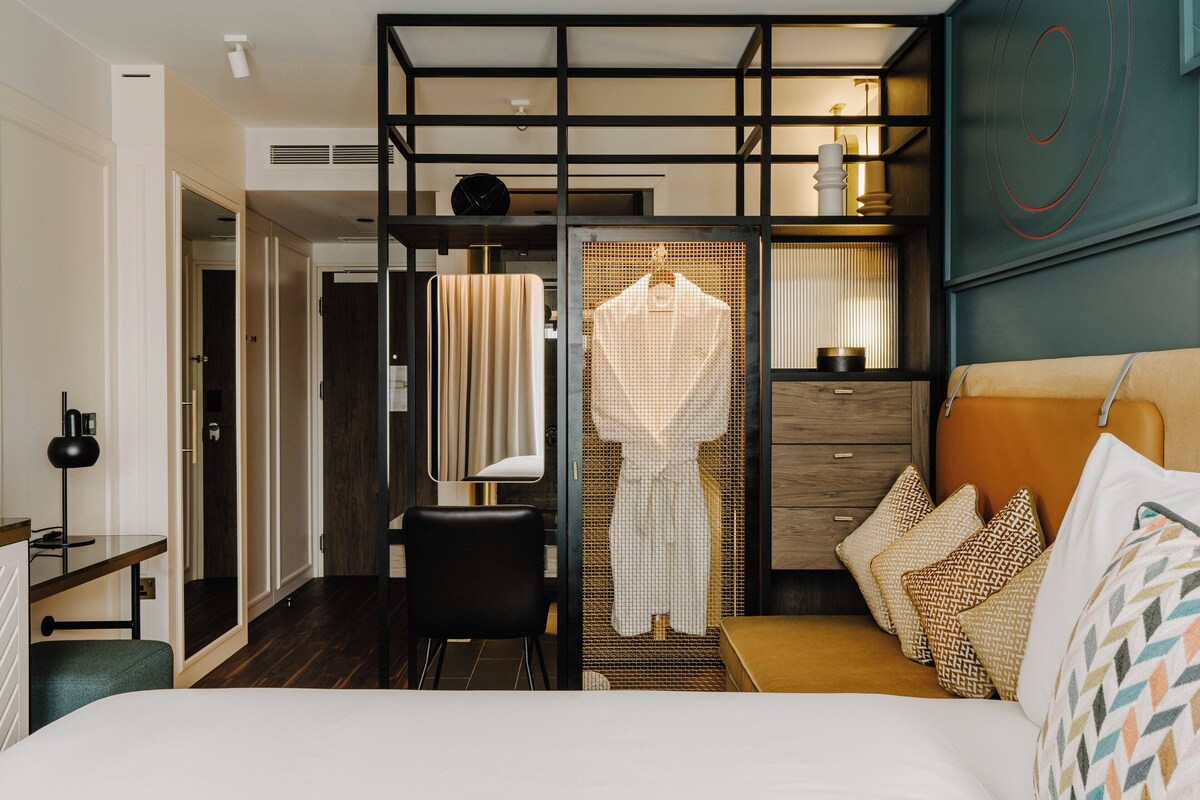 A hotel room with a bed, a desk, a chair, a wardrobe, and a hanging garment.