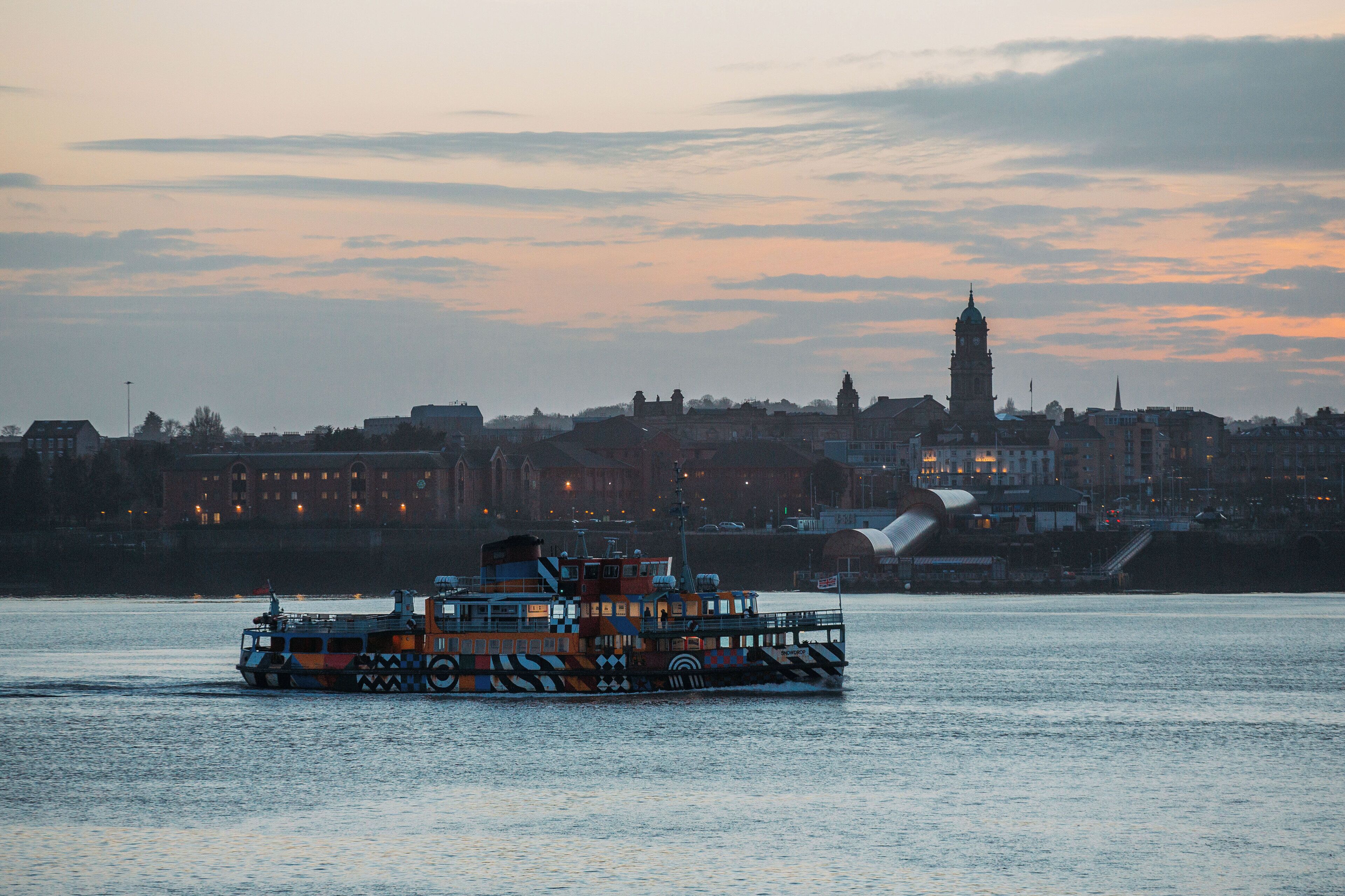 Een rivierboot met een kleurrijk ontwerp, en op de achtergrond een stadszicht met gebouwen en een klokkentoren.