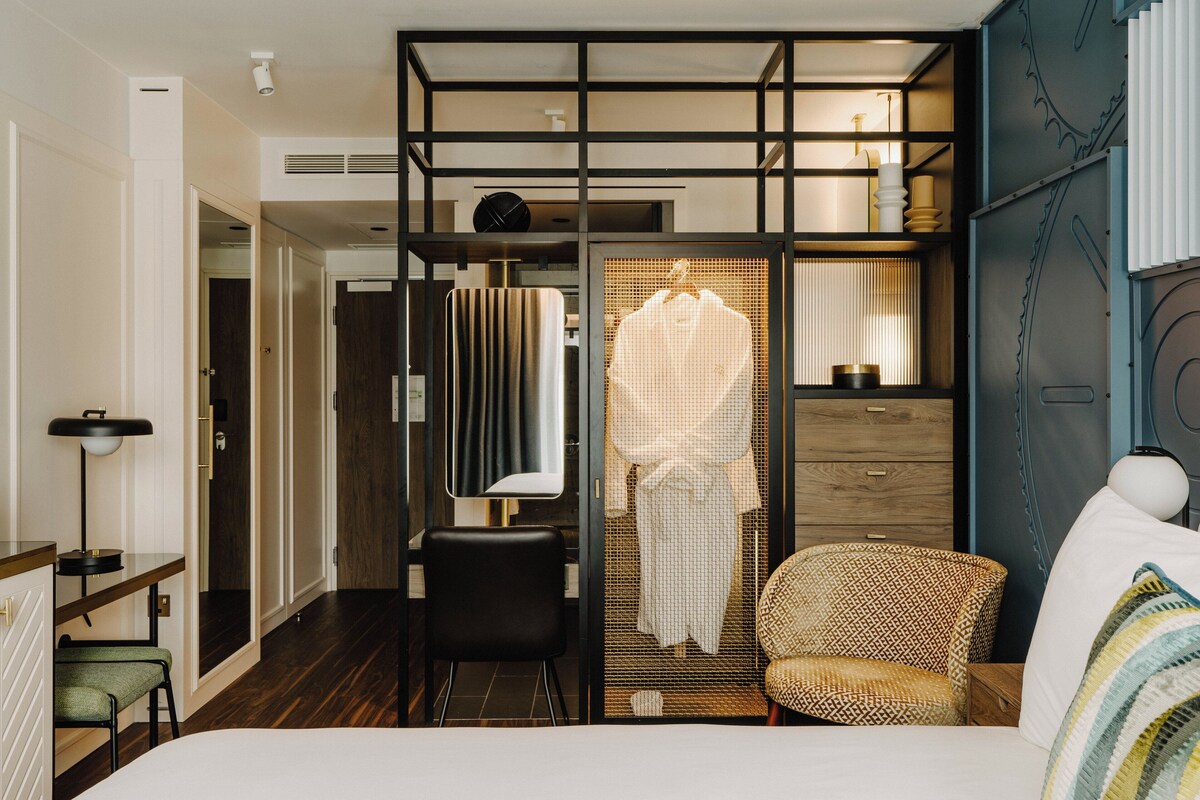 A modern hotel room with a bed, a desk, a chair, and a wardrobe.