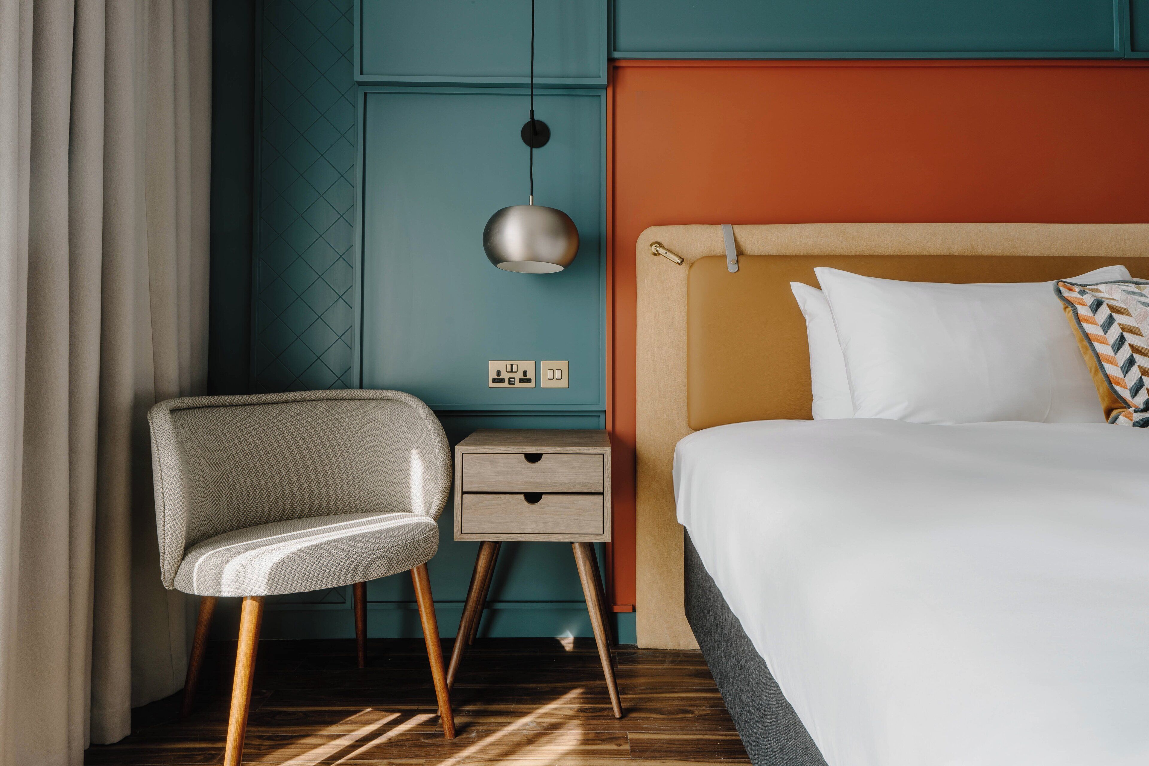 Quarto de hotel com uma cama, mesa de cabeceira, cadeira e luminária fixada na parede.