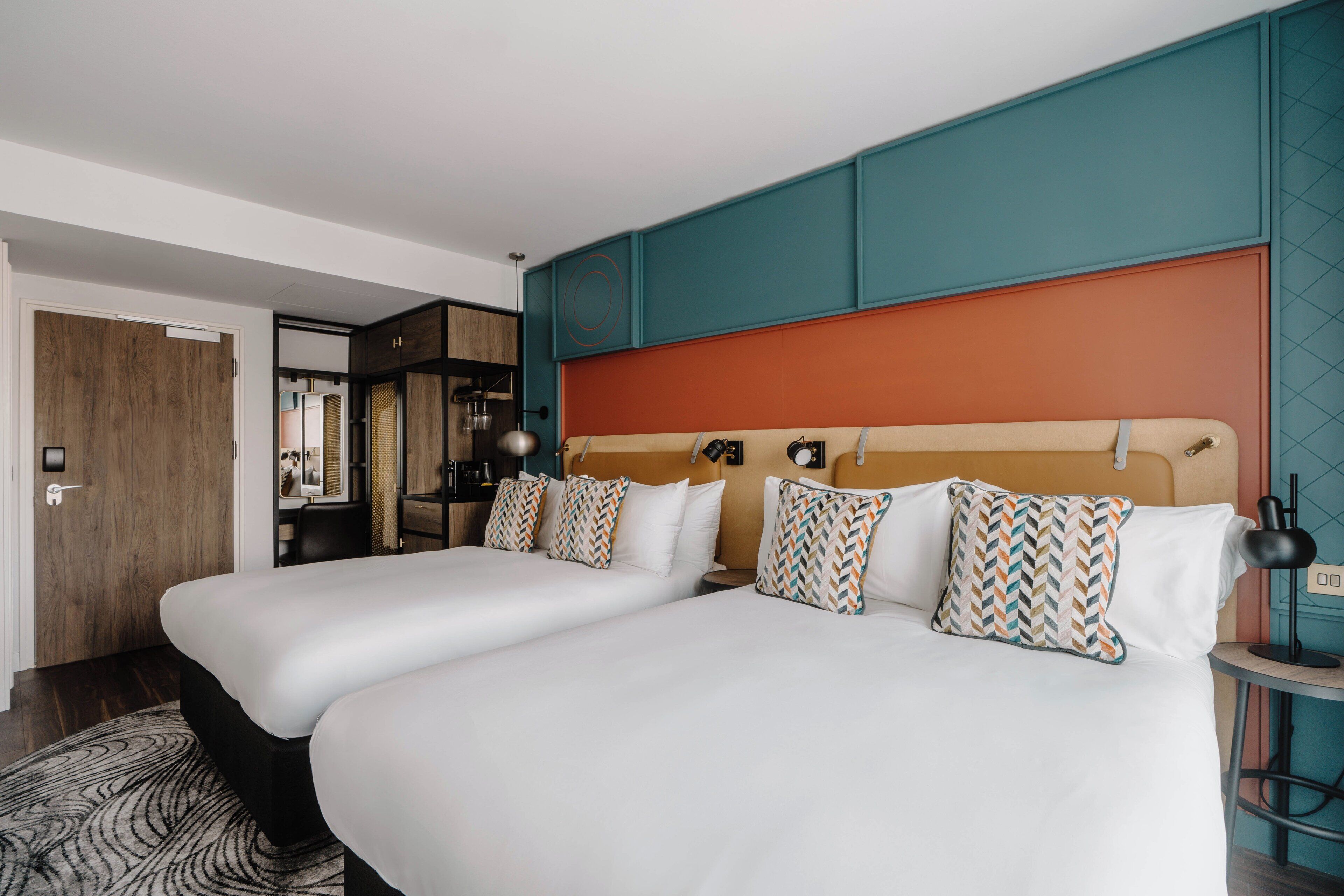 A hotel room with two beds, a wooden door, a kitchenette, and a view of the city.