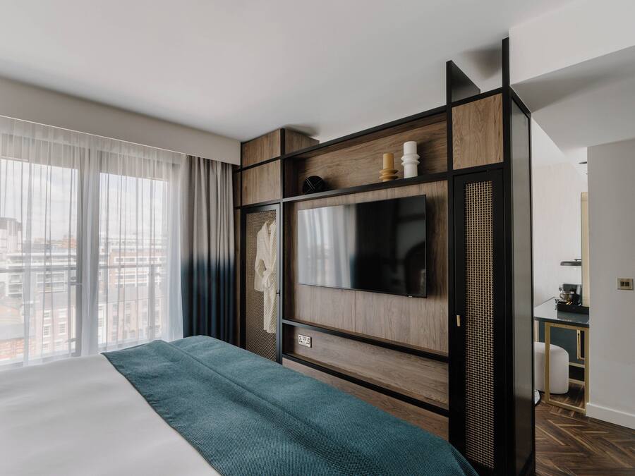 A modern hotel room with a large bed, a TV mounted on a wooden wall, and a view of the city through large windows.