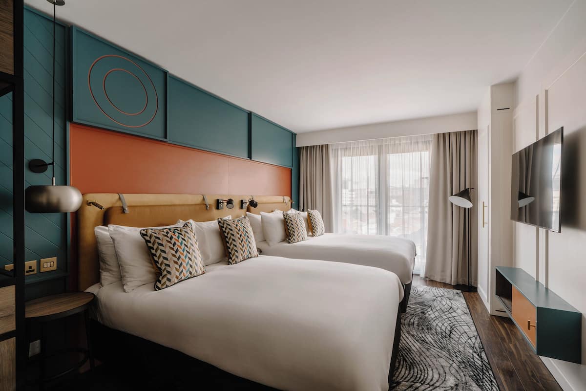 A hotel room with two beds, a TV, a desk, and a large window with curtains.